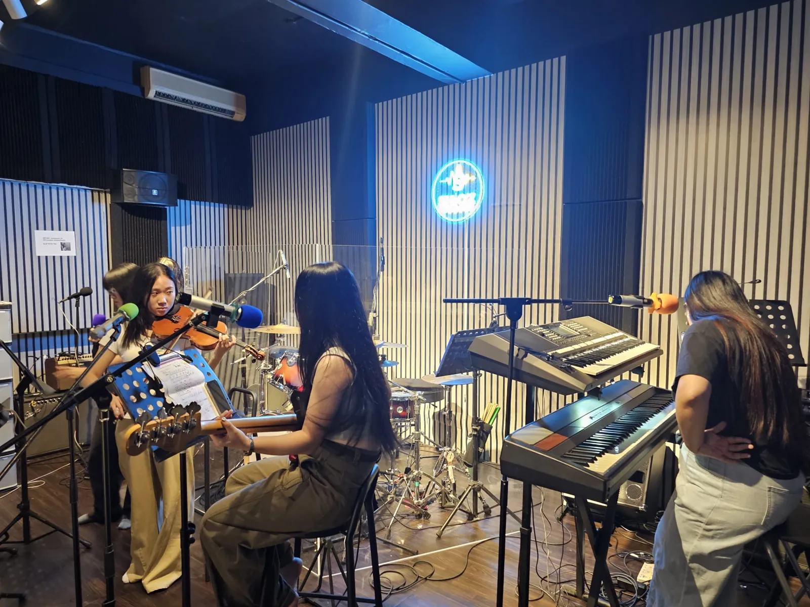 Musicians rehearsing with keyboards and violin under the neon logo